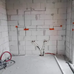 Surowy stan instalacji w pomieszczeniu z bloków: widoczne przewody elektryczne w peszlach, puszki instalacyjne, rury wodne z izolacją termiczną, instalacja kanalizacyjna przy podłodze.