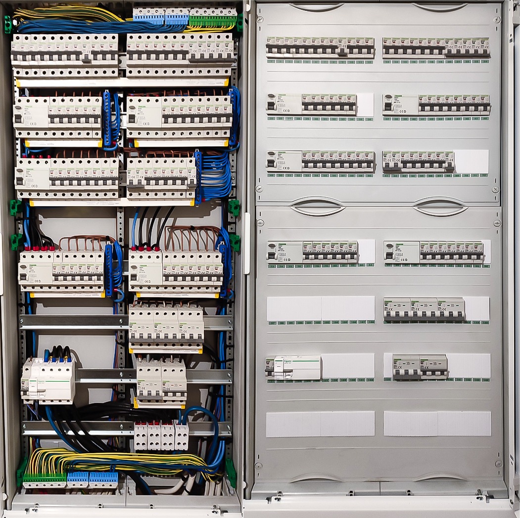 Otwarta szafa rozdzielcza z widocznymi bezpiecznikami, przewodami i elementami instalacji elektrycznej, uporządkowane okablowanie w kolorach niebieskim, żółtym, zielonym i czarnym.