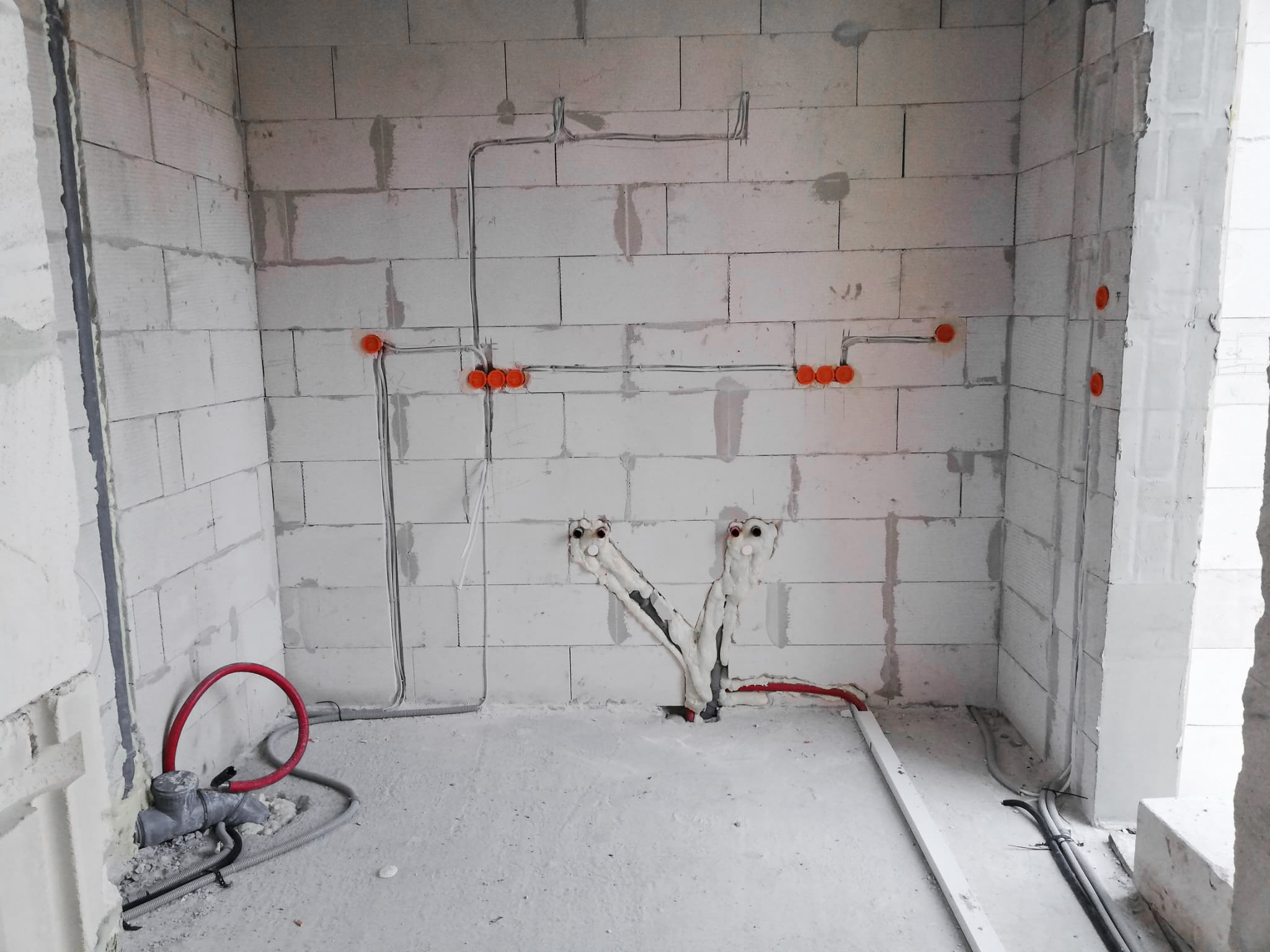 Surowy stan instalacji w pomieszczeniu z bloków: widoczne przewody elektryczne w peszlach, puszki instalacyjne, rury wodne z izolacją termiczną, instalacja kanalizacyjna przy podłodze.
