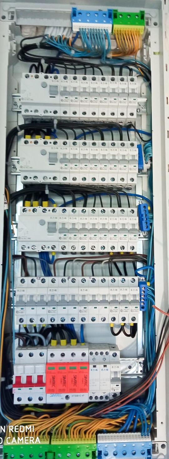 Rozdzielnica elektryczna z zainstalowanymi bezpiecznikami, przewodami o różnych kolorach i listwami zaciskowymi, widoczne zabezpieczenia przeciwprzepięciowe, zdjęcie zrobione z bliska.