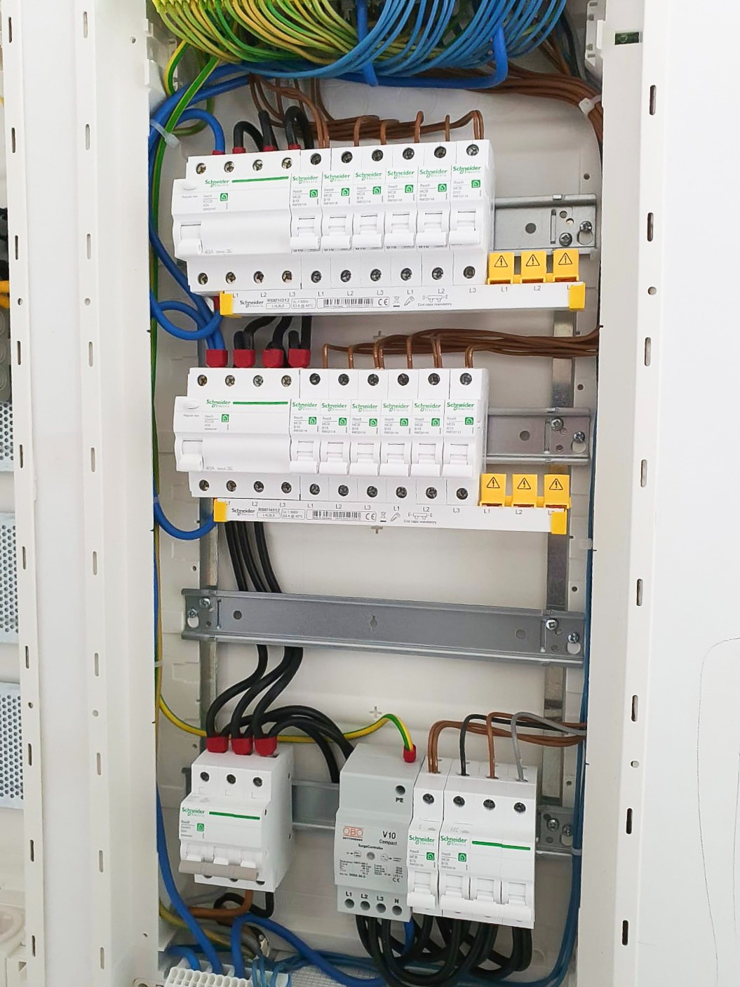 Wnętrze skrzynki elektrycznej z zainstalowanymi zabezpieczeniami przepięciowymi Schneider, przewodami w różnych kolorach i listwami zaciskowymi, widoczne żółte oznaczenia faz.