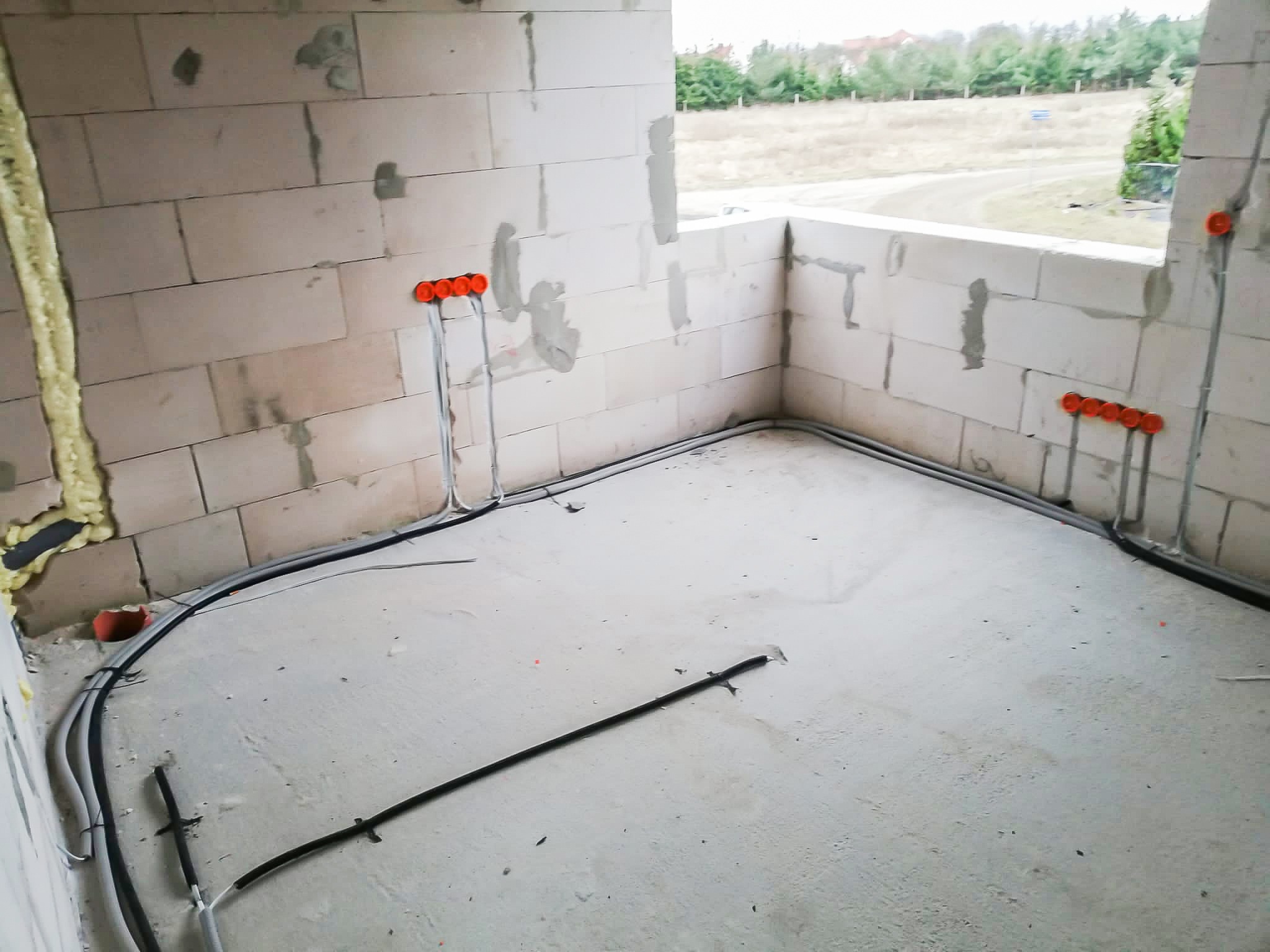 Wnętrze w surowym stanie z widocznymi przewodami elektrycznymi poprowadzonymi wzdłuż ścian i podłogi, pomarańczowe puszki instalacyjne zamontowane w ścianach z betonu komórkowego, pianka montażowa...