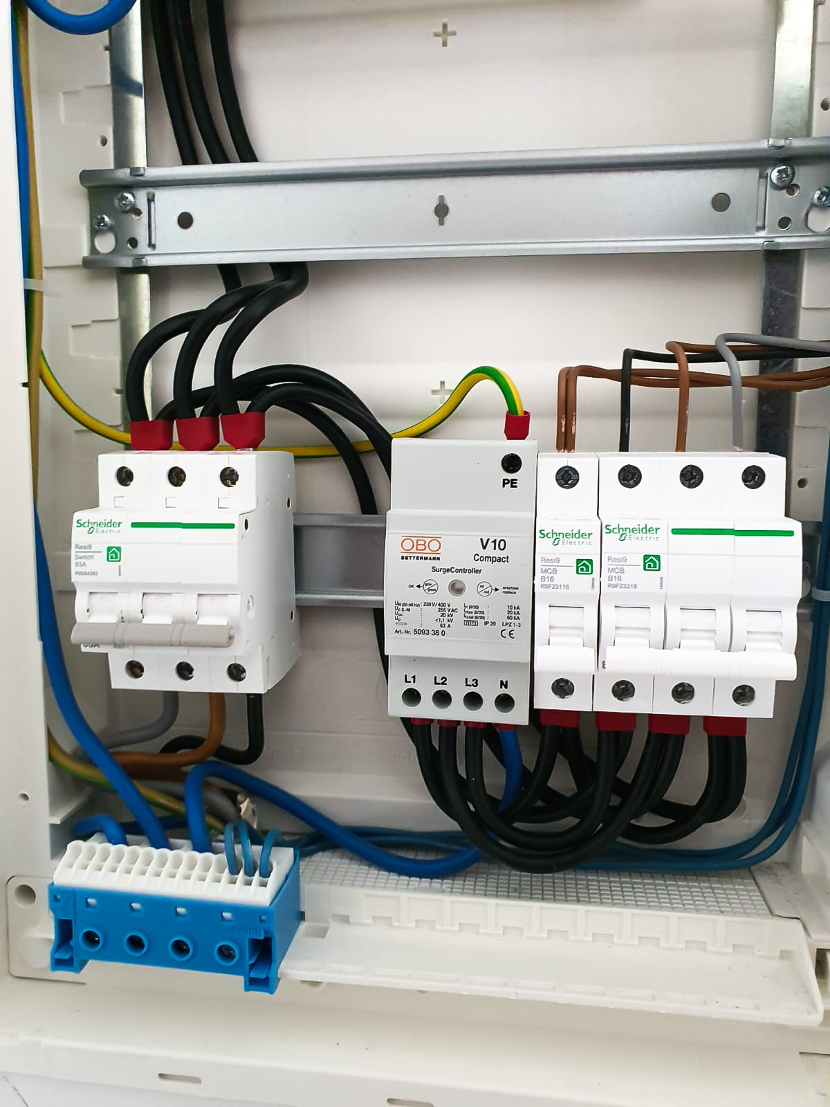 Rozdzielnica elektryczna z zamontowanymi zabezpieczeniami przepięciowymi OBO V10 i wyłącznikami nadprądowymi Schneider Electric Resi9, widoczne przewody i listwa montażowa.