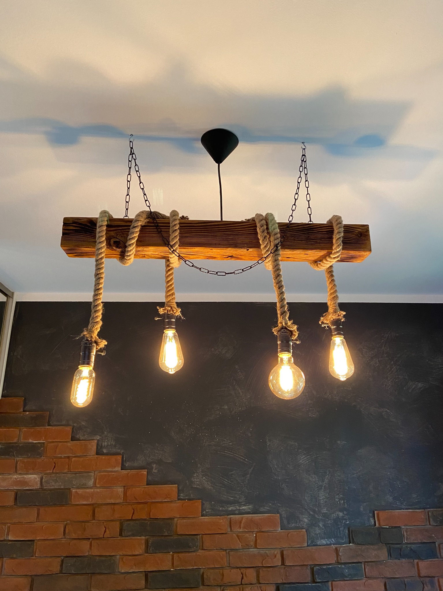 Rustykalna lampa wisząca z drewnianej belki, sznura jutowego i czterech żarówek Edisona, zawieszona na łańcuchach, na tle ściany z cegieł i czarnej ściany z fakturą.