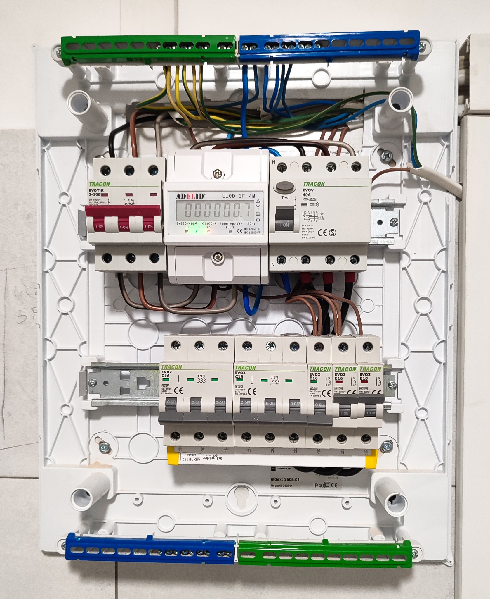 Rozdzielnica elektryczna z zainstalowanymi bezpiecznikami, licznikiem energii i przewodami w kolorach niebieskim, brązowym, żółto-zielonym; widoczne marki Tracon i Adelid.