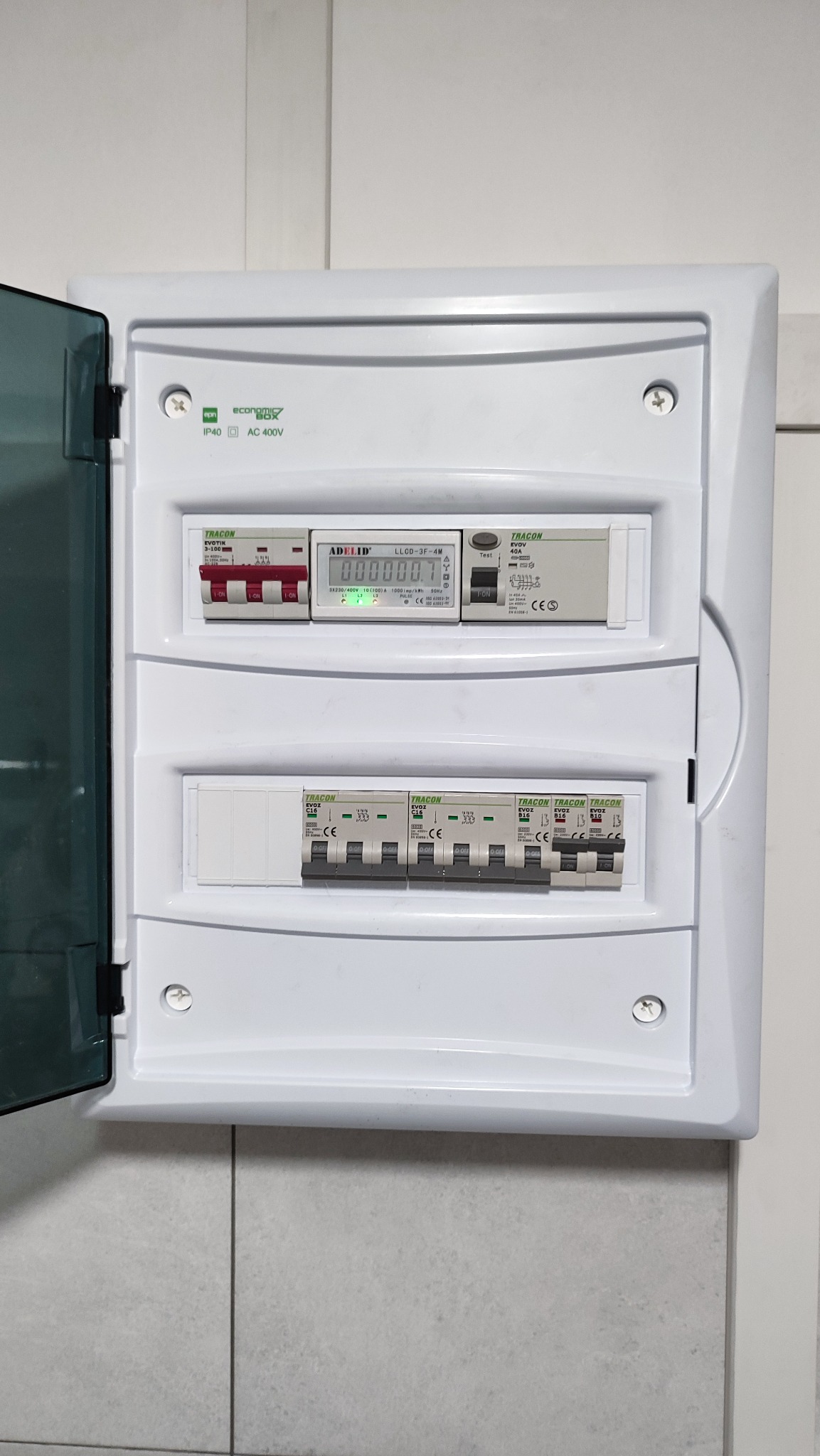 Otwarta skrzynka elektryczna z zainstalowanymi bezpiecznikami i licznikiem energii, widoczne oznaczenia producentów Tracon i Adelid, zamontowana na jasnej ścianie.