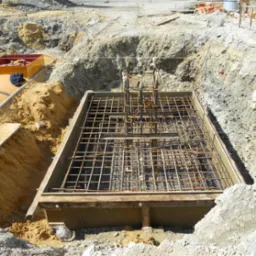 Zbrojenie fundamentu w wykopie, szalunek z desek, widoczne pręty stalowe tworzące siatkę. W tle ziemia i materiały budowlane. Jasne światło dzienne.