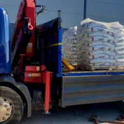 Niebieska ciężarówka z czerwoną platformą załadunkową i dźwigiem, przewożąca paletę z workami z zaprawą murarską marki Karix, na tle szarego budynku przemysłowego.