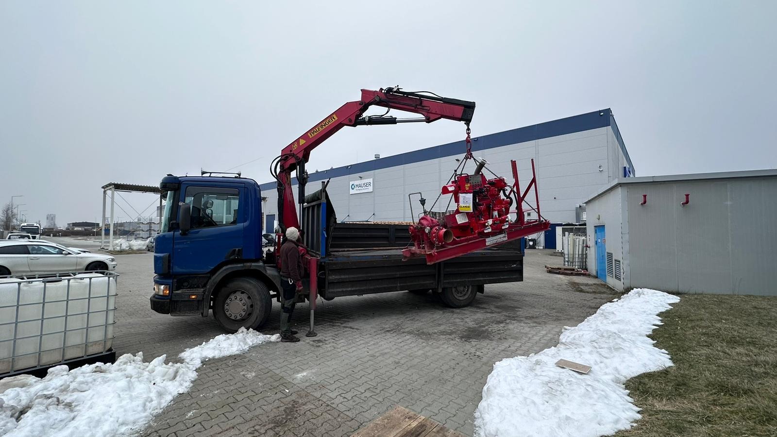 Niebieska ciężarówka z dźwigiem Palfinger podnosi czerwoną pompę na skrzynię ładunkową, pracownik w kurtce obserwuje proces, w tle budynek przemysłowy Mauser i resztki śniegu.