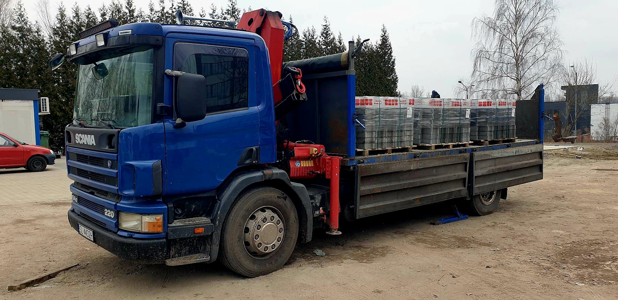 Niebieska ciężarówka Scania z dźwigiem HDS przewożąca palety z kostką brukową na skrzyni ładunkowej, widok z boku na szarym tle.