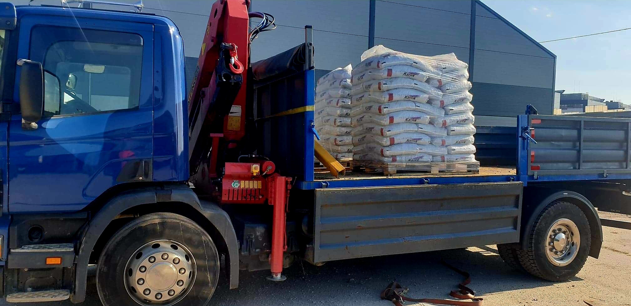 Niebieska ciężarówka z czerwoną platformą załadunkową i dźwigiem, przewożąca paletę z workami z zaprawą murarską marki Karix, na tle szarego budynku przemysłowego.