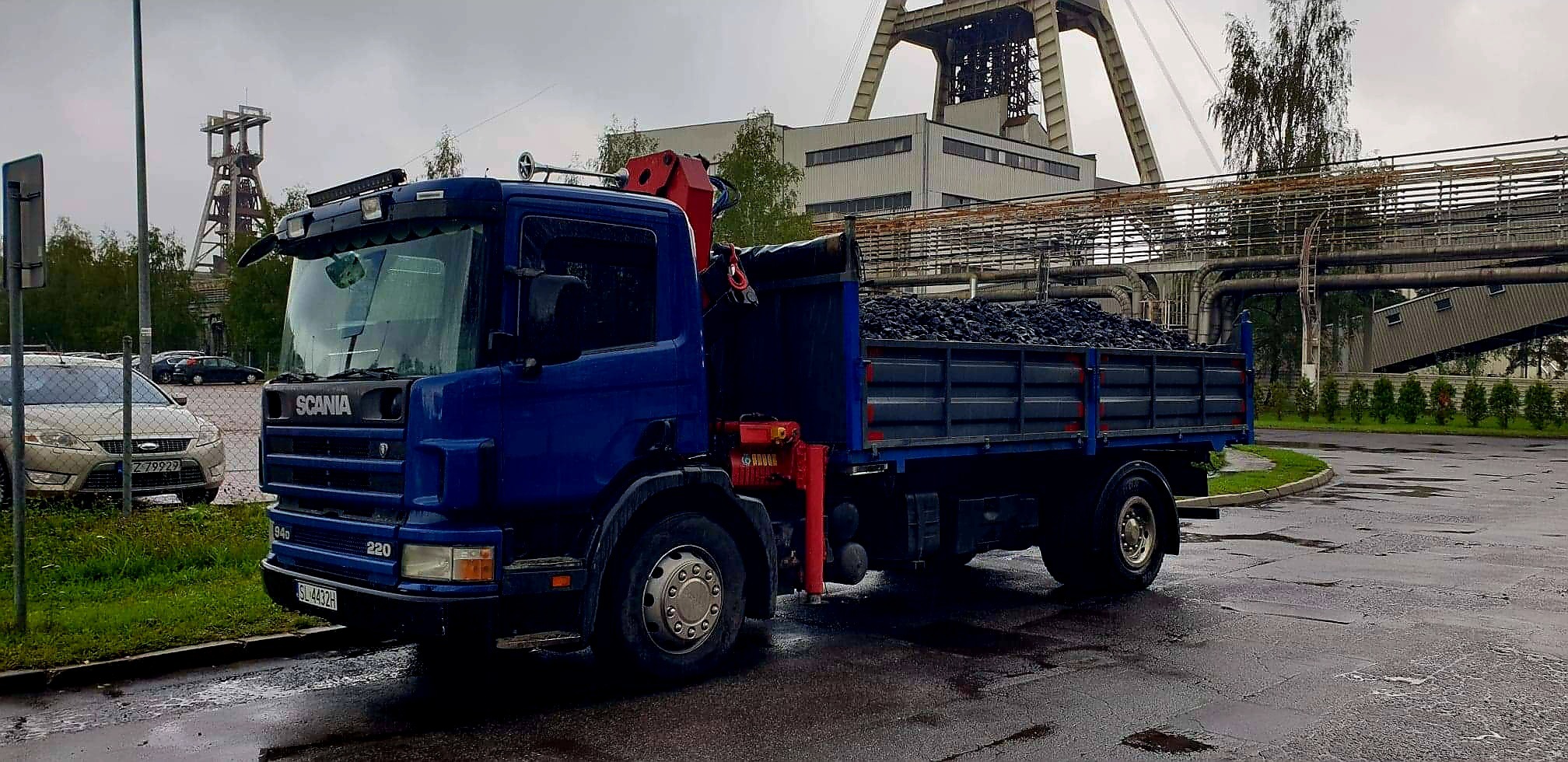 Niebieska ciężarówka Scania z czerwonym żurawiem załadowana węglem, na tle przemysłowego krajobrazu z szybem kopalnianym i rurociągami.