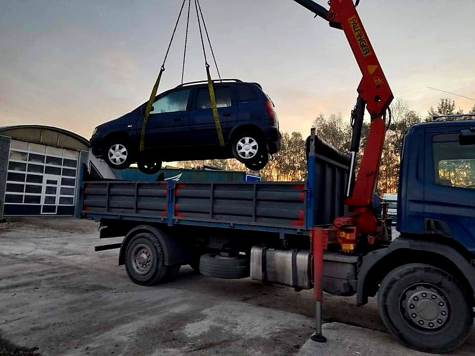 Ciemnoniebieski samochód osobowy podnoszony dźwigiem zamontowanym na niebieskiej ciężarówce z otwartą skrzynią ładunkową na tle szarego nieba i drzew.