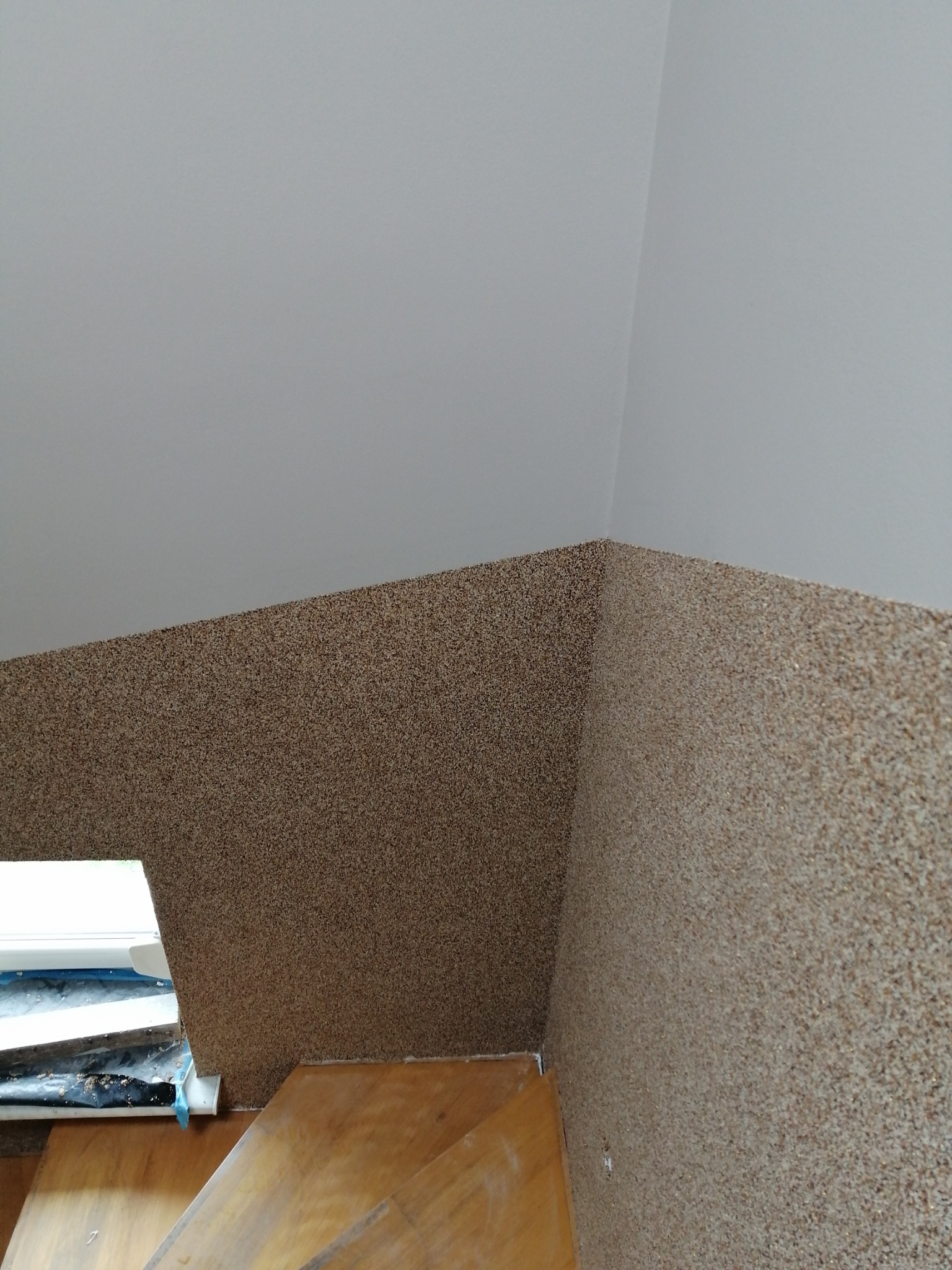 Tekstura ściany pokryta dekoracyjnym tynkiem mozaikowym w odcieniach beżu i brązu, widoczna przy schodach i oknie, z zabezpieczeniem folią malarską.