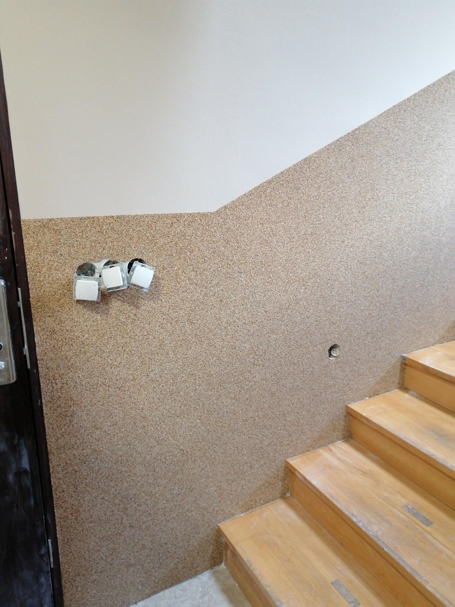 Ściana klatki schodowej wykończona tynkiem mozaikowym w odcieniach beżu i brązu, z widocznymi trzema gniazdkami elektrycznymi i fragmentem drewnianych schodów.