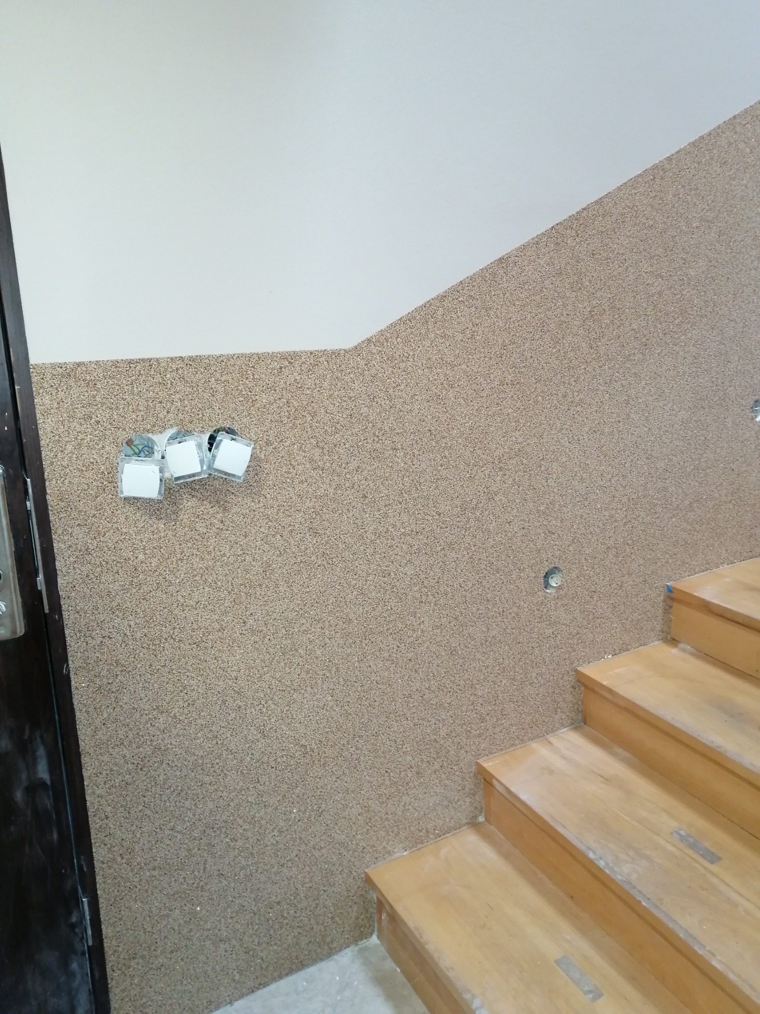 Ściana pokryta dekoracyjnym tynkiem mozaikowym w odcieniach beżu i brązu, widoczne puszki elektryczne i włączniki światła, drewniane schody w tle.