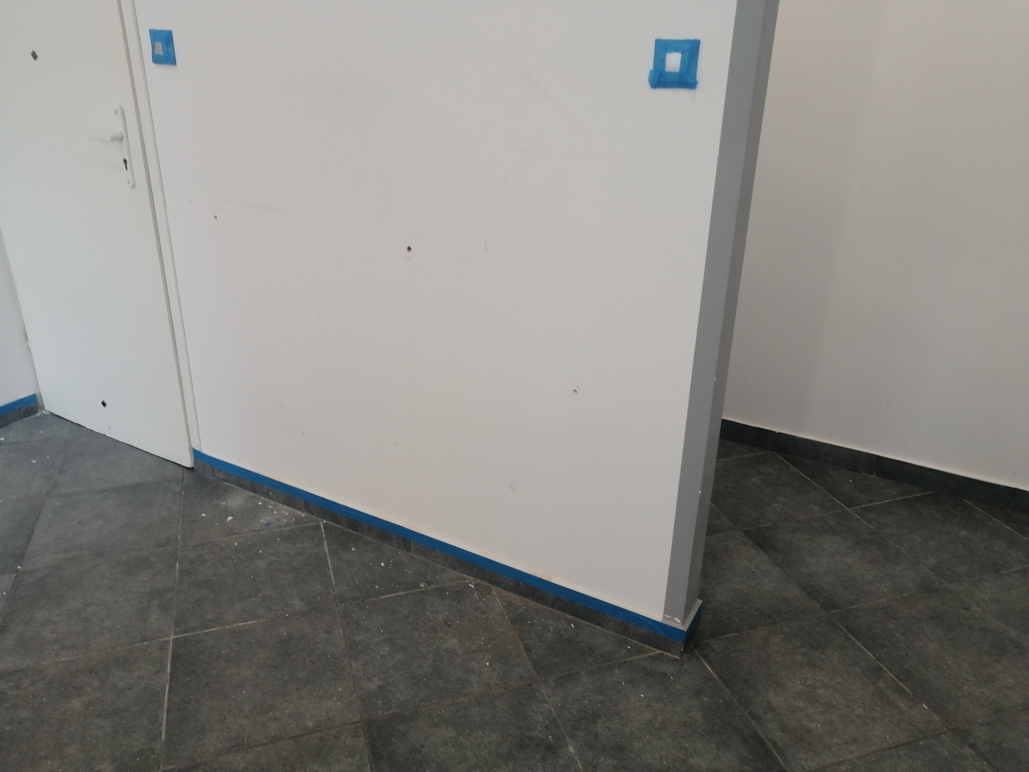 Przygotowanie do malowania: ściana i drzwi zabezpieczone niebieską taśmą malarską, widoczne drobne zabrudzenia na podłodze z płytek.