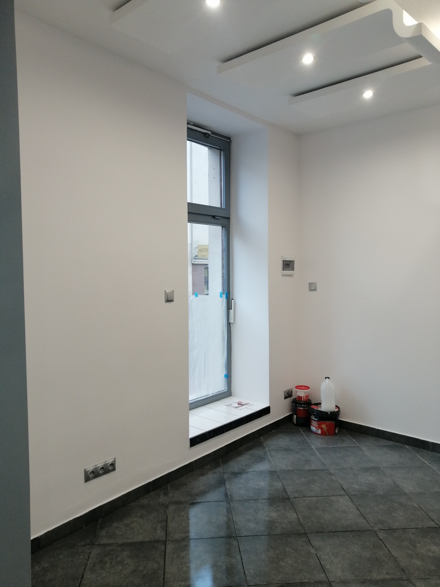 Wnętrze pokoju po remoncie, świeżo pomalowane ściany na biało, widoczne puszki z farbą i akcesoria malarskie przy ścianie, ciemna podłoga z płytek, okno zabezpieczone folią malarską.