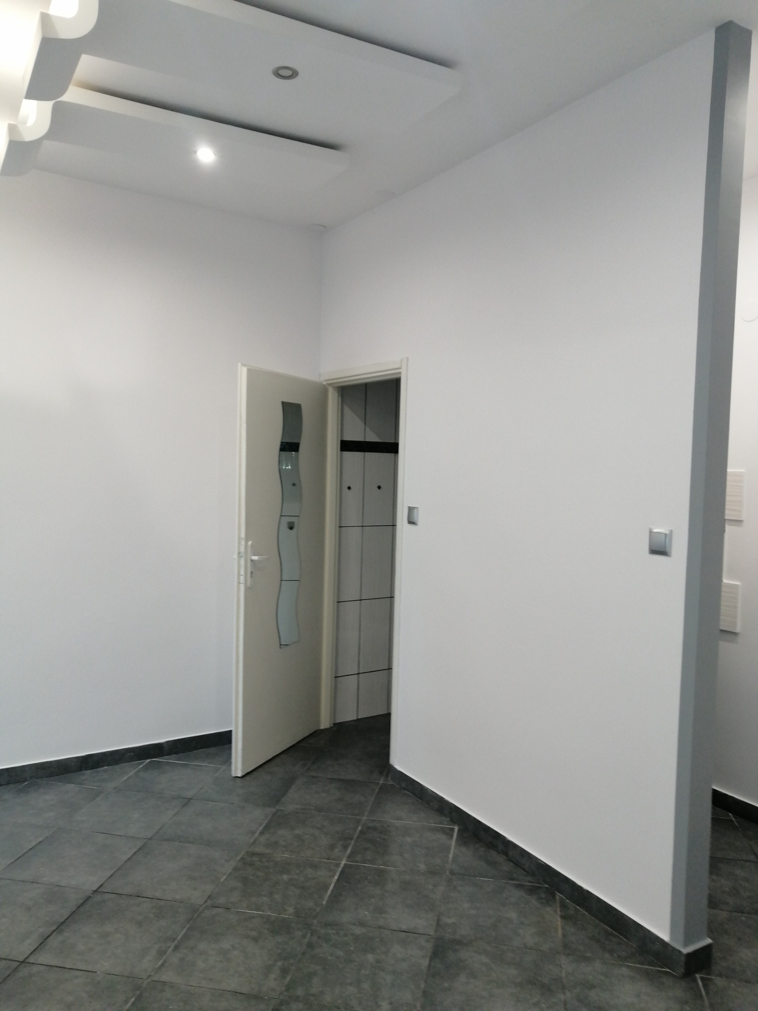 Wnętrze pomalowanego na biało pokoju z ciemnoszarą podłogą z płytek, widoczne otwarte drzwi do łazienki z białymi płytkami.