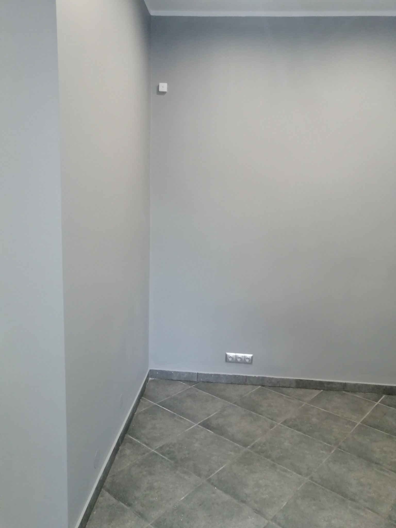 Świeżo pomalowane szare ściany w pokoju z ciemnoszarą podłogą z płytek ułożonych w karo, widoczne gniazdka elektryczne i włącznik światła.