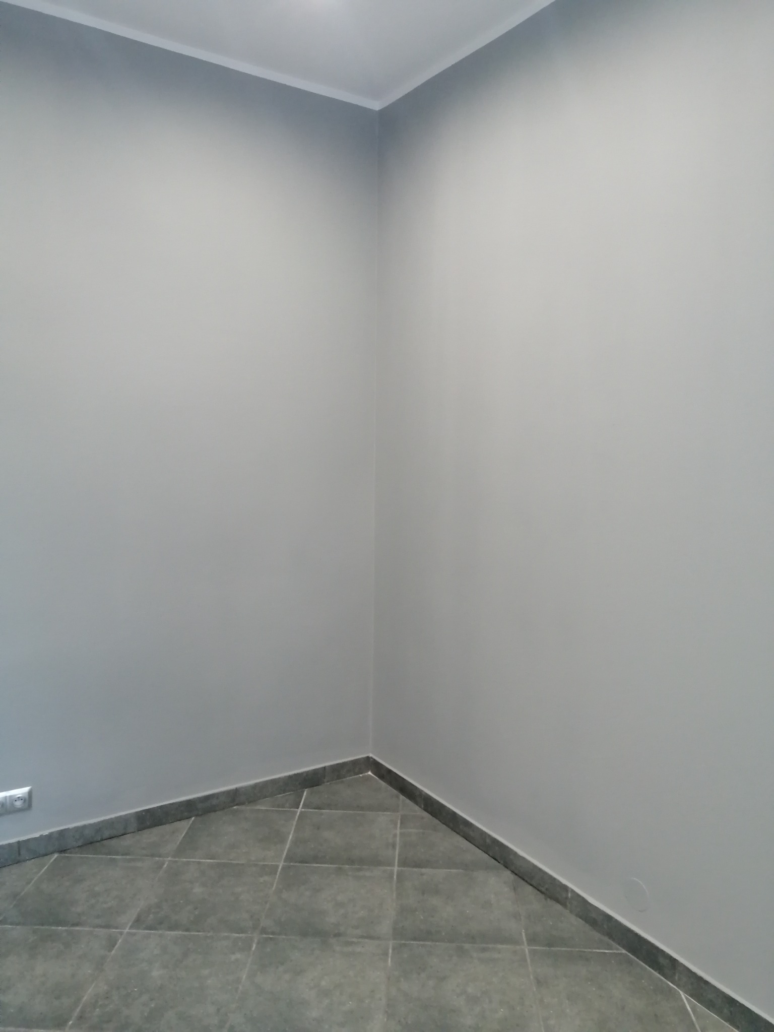 Pomalowane na szaro wnętrze pokoju z szarą podłogą z płytek i listwami przypodłogowymi. Widoczne gniazdko elektryczne po lewej stronie.