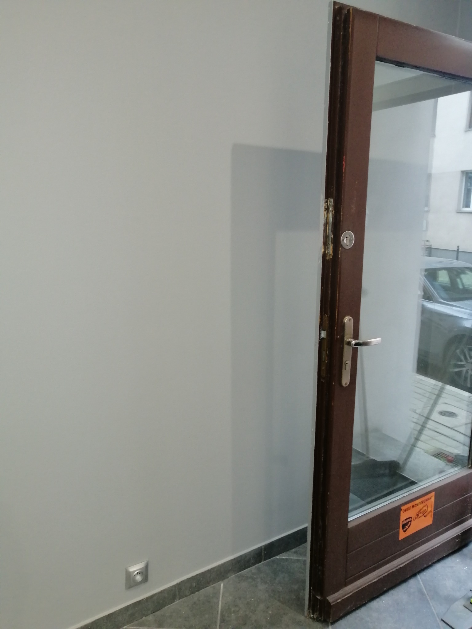 Szara ściana świeżo po malowaniu, widoczna brązowa rama drzwi z szybą z naklejką ostrzegawczą, szara podłoga z listwą przypodłogową, gniazdko elektryczne na ścianie.