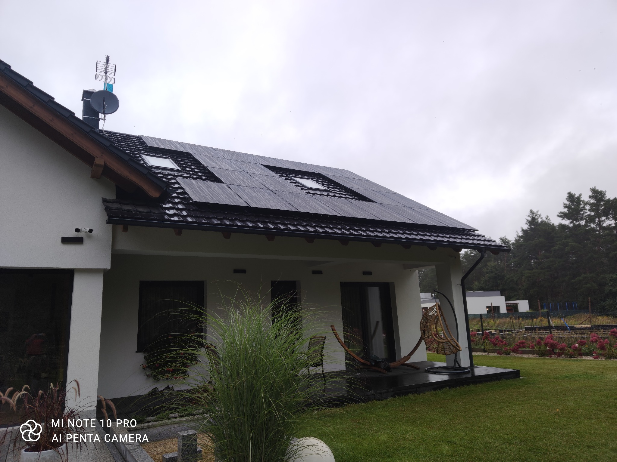 Czarny dach z panelami słonecznymi i oknami dachowymi na nowoczesnym białym domu z ogrodem, antena satelitarna na kominie.