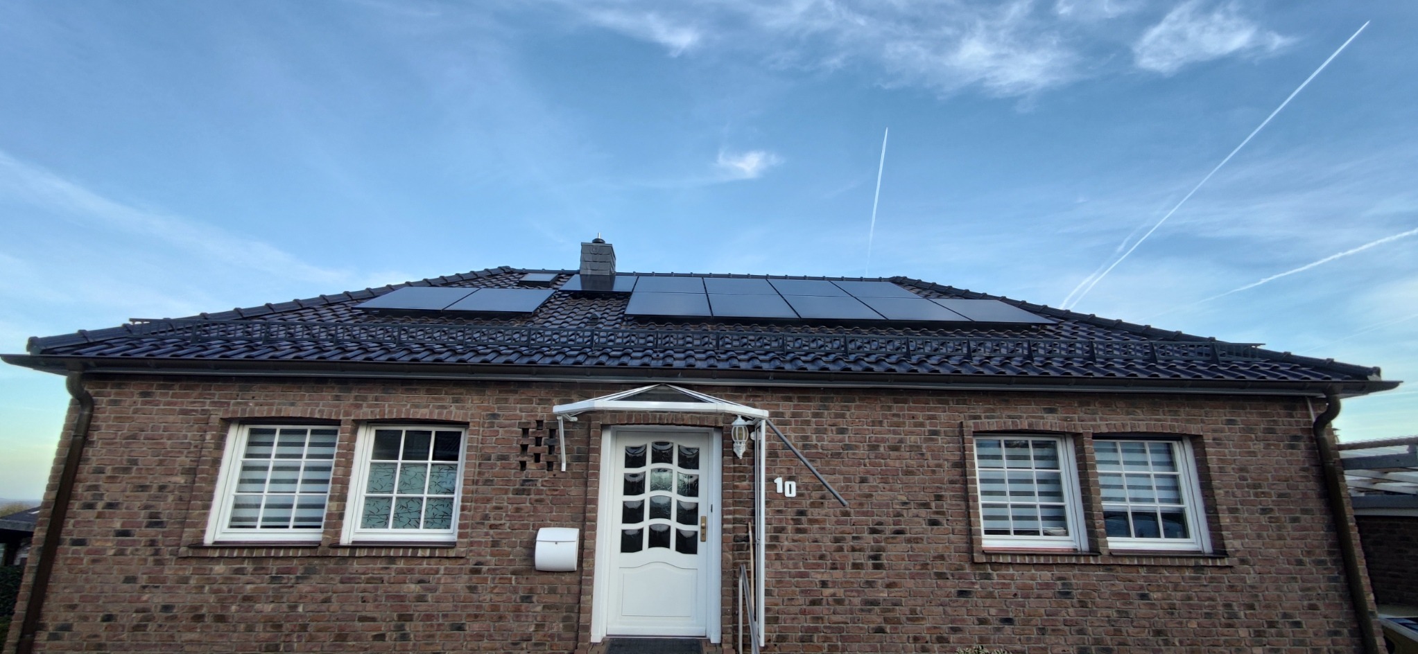 Dom z cegły z panelami słonecznymi na dachu i smugami kondensacyjnymi na niebie.