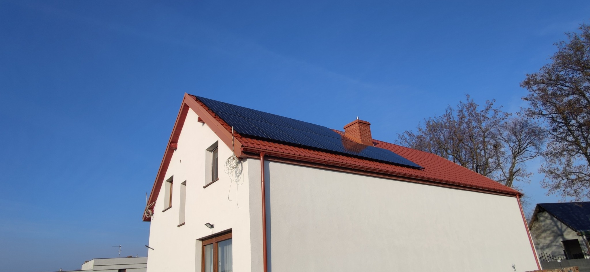 Biały dom jednorodzinny z panelami słonecznymi na czerwonym dachu, widoczne okna, komin i fragment sąsiedniego budynku w słoneczny dzień.