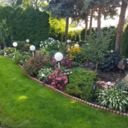 Estetyczny ogród z równo przystrzyżonym trawnikiem, ozdobnymi krzewami, kwiatami i białymi kulistymi lampami. Brzeg trawnika wykończony drewnianymi palisadami.