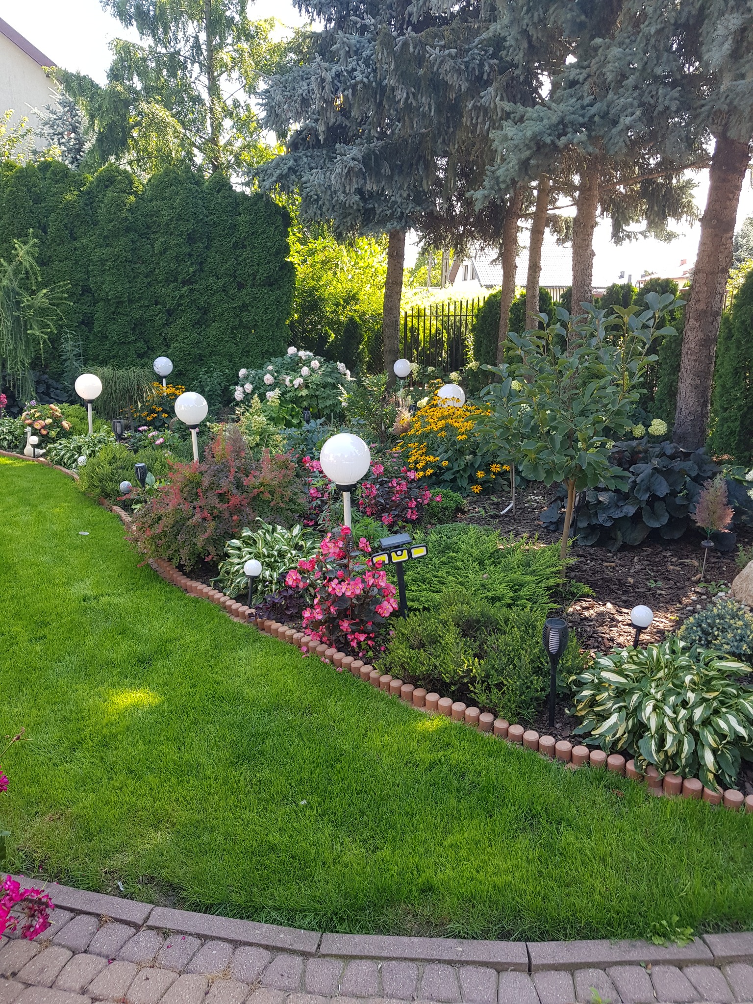 Estetyczny ogród z równo przystrzyżonym trawnikiem, ozdobnymi krzewami, kwiatami i białymi kulistymi lampami. Brzeg trawnika wykończony drewnianymi palisadami.