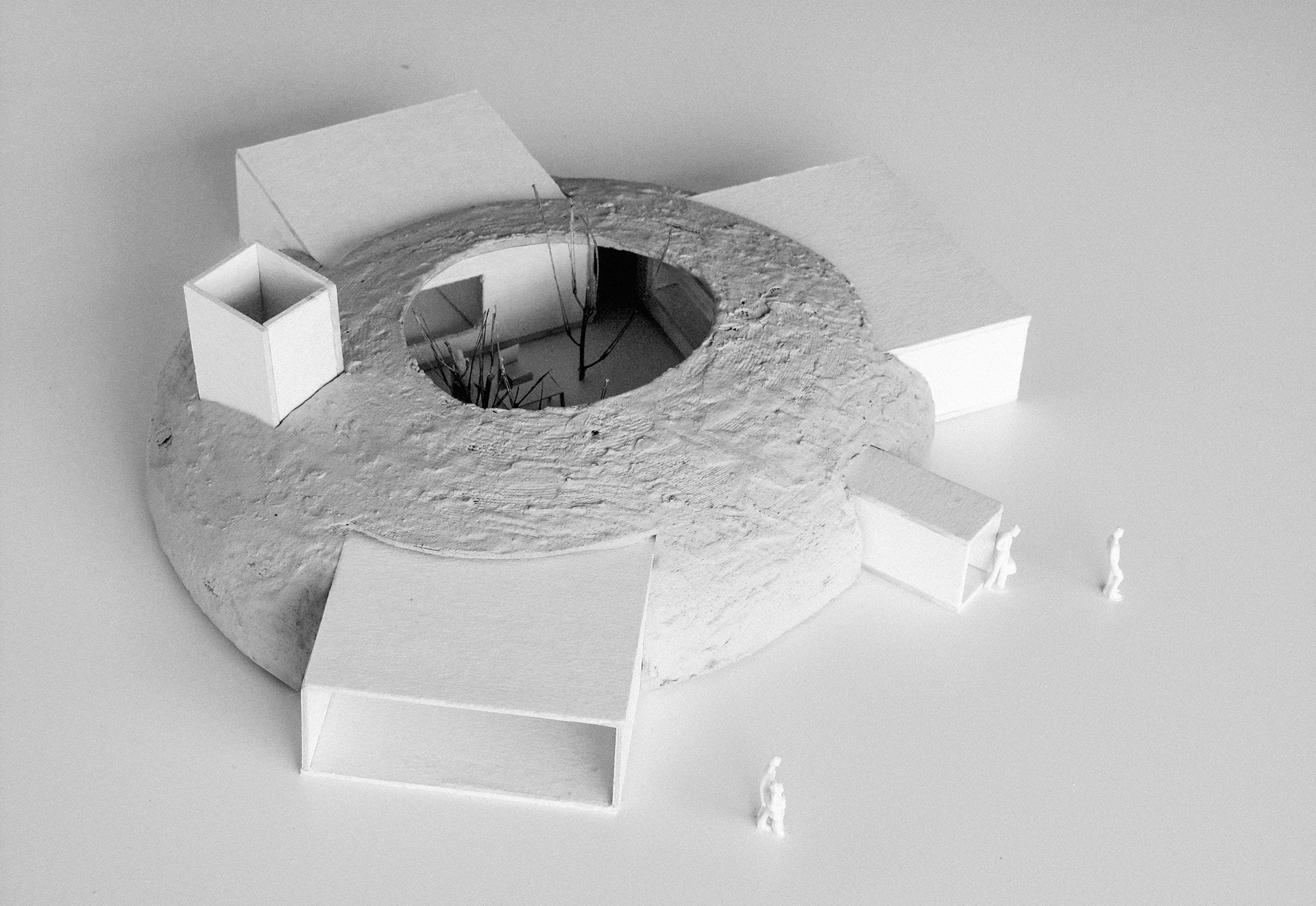 Czarno-biały model architektoniczny budynku z okrągłym dziedzińcem i dołączonymi prostopadłościennymi bryłami, uzupełniony o miniaturowe figurki ludzi.