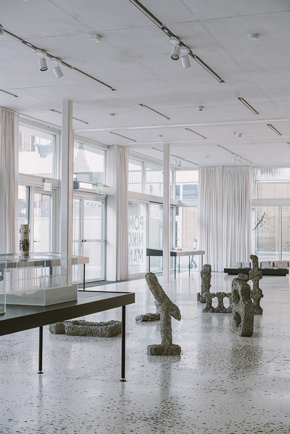 Jasne, przestronne wnętrze budynku usługowego z dużymi oknami, białymi zasłonami i betonową podłogą w lastryko, prezentujące ekspozycję rzeźb z szarego betonu.
