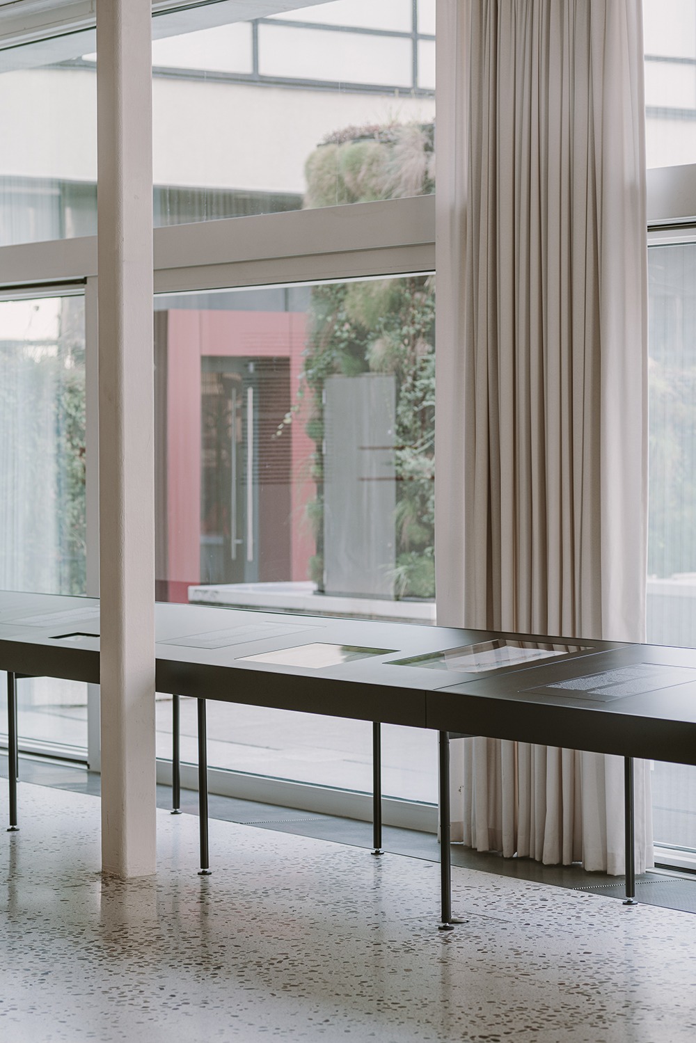 Wnętrze biura z długim stołem ekspozycyjnym na cienkich nogach, z wbudowanymi gablotami, na tle okna z widokiem na zieloną ścianę i budynek z czerwonymi elementami.