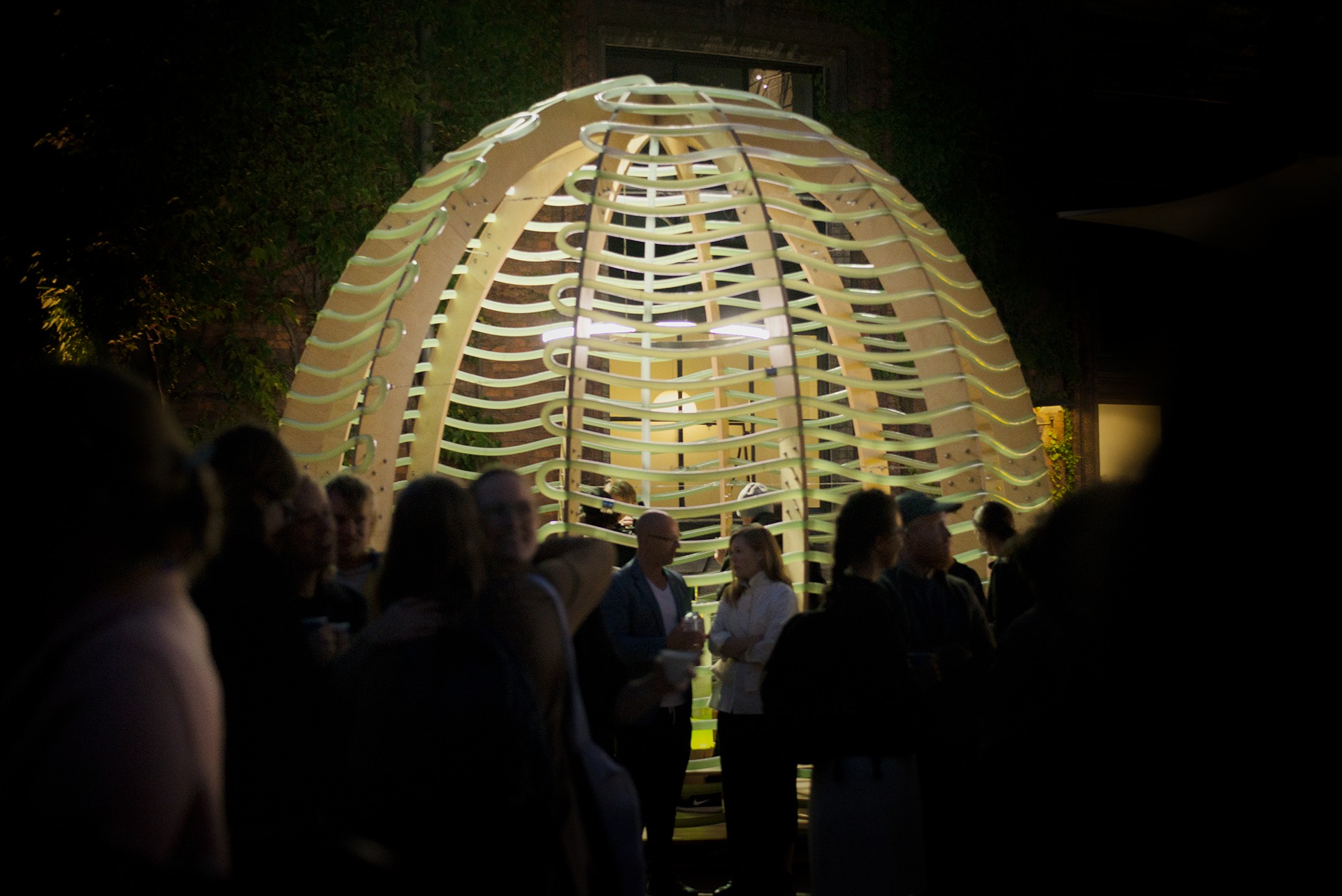 Oświetlona, ażurowa konstrukcja kopułowa z drewna i tworzywa sztucznego na tle ciemnego otoczenia z zielenią, otoczona przez grupę ludzi podczas wieczornego wydarzenia.