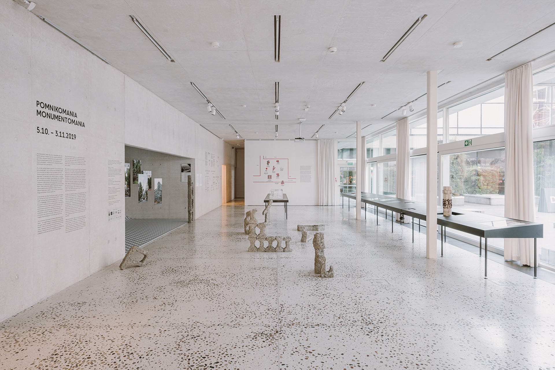 Przestronne wnętrze galerii z betonową posadzką lastryko, prezentujące ekspozycję rzeźb z betonu, długie stoły ekspozycyjne i duże okna z białymi zasłonami.