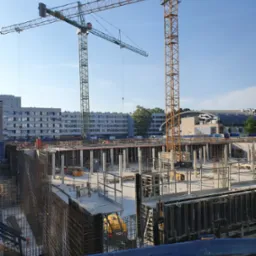 Rozległy plac budowy z dwoma żurawiami wieżowymi, widoczne zbrojenia i szalunki fundamentów, betonowe kolumny, w tle budynki mieszkalne i hala.