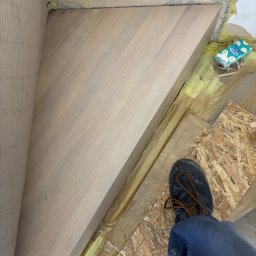 Wiktor Lewandowicz Wooden Solutions - Drewniane schody w trakcie montażu, widok z góry na stopień z fragmentem buta pracownika. Żółta siatka zabezpieczająca na ścianie i folia ochronna na podłodze.