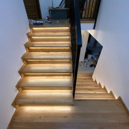 Wiktor Lewandowicz Wooden Solutions - Nowoczesne schody z jasnego drewna z podświetleniem LED, widok z góry. Minimalistyczna balustrada z czarnych prętów. Białe ściany. Widoczne elementy wykończenia wnętrza.