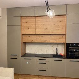 Wiktor Lewandowicz Wooden Solutions - Nowoczesna kuchnia z szarymi szafkami i drewnianymi akcentami, czarnym blatem i marmurowym backsplash. Widoczny fragment sofy i osoba w tle. Oświetlenie sufitowe.