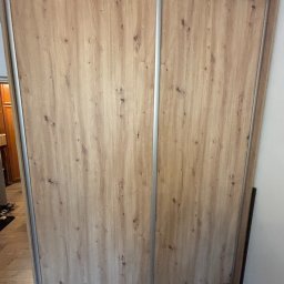 Wiktor Lewandowicz Wooden Solutions - Zamknięta szafa przesuwna z drewnopodobnymi frontami i srebrnymi obramowaniami, widoczna z bliska, z fragmentem wnętrza pomieszczenia po lewej stronie.