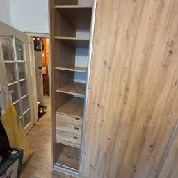 Wiktor Lewandowicz Wooden Solutions - Jasna, drewnopodobna szafa wnękowa z przesuwnymi drzwiami i otwartymi półkami, widoczna z boku, z perspektywy podłogi. W tle drzwi do innego pomieszczenia.