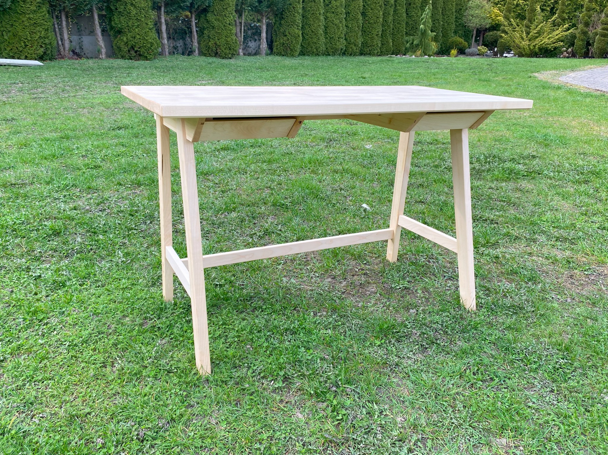 Surowy, jasny, drewniany stół z dwiema szufladami, stojący na trawie w ogrodzie, widoczne niedoskonałości obróbki drewna na nogach