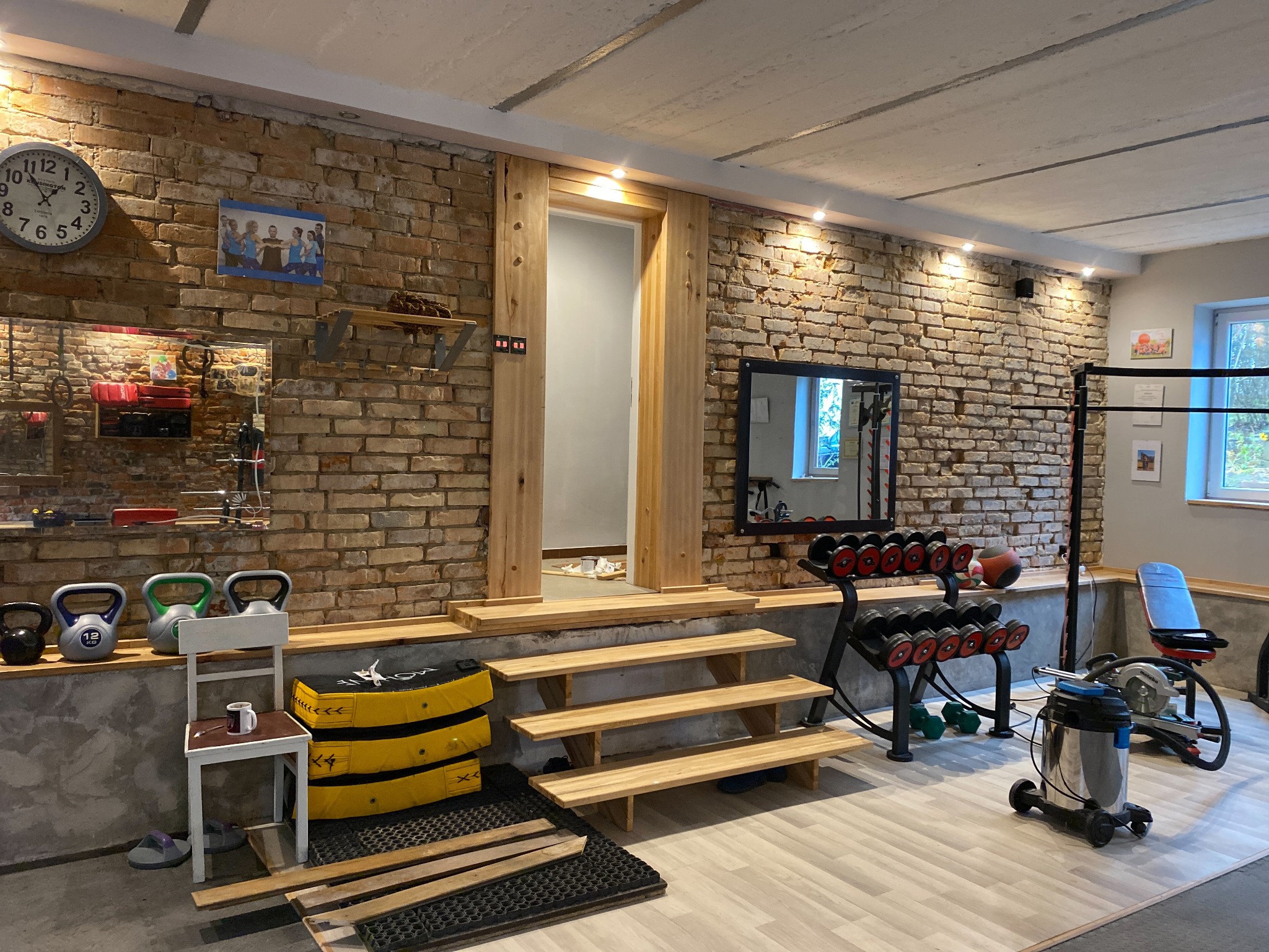 Domowa siłownia z ceglaną ścianą, drewnianymi stopniami prowadzącymi do drzwi, sprzętem do ćwiczeń i narzędziami, widoczne kettlebells, hantle, piłka lekarska, drabinki, ławka do ćwiczeń, odkurzacz...