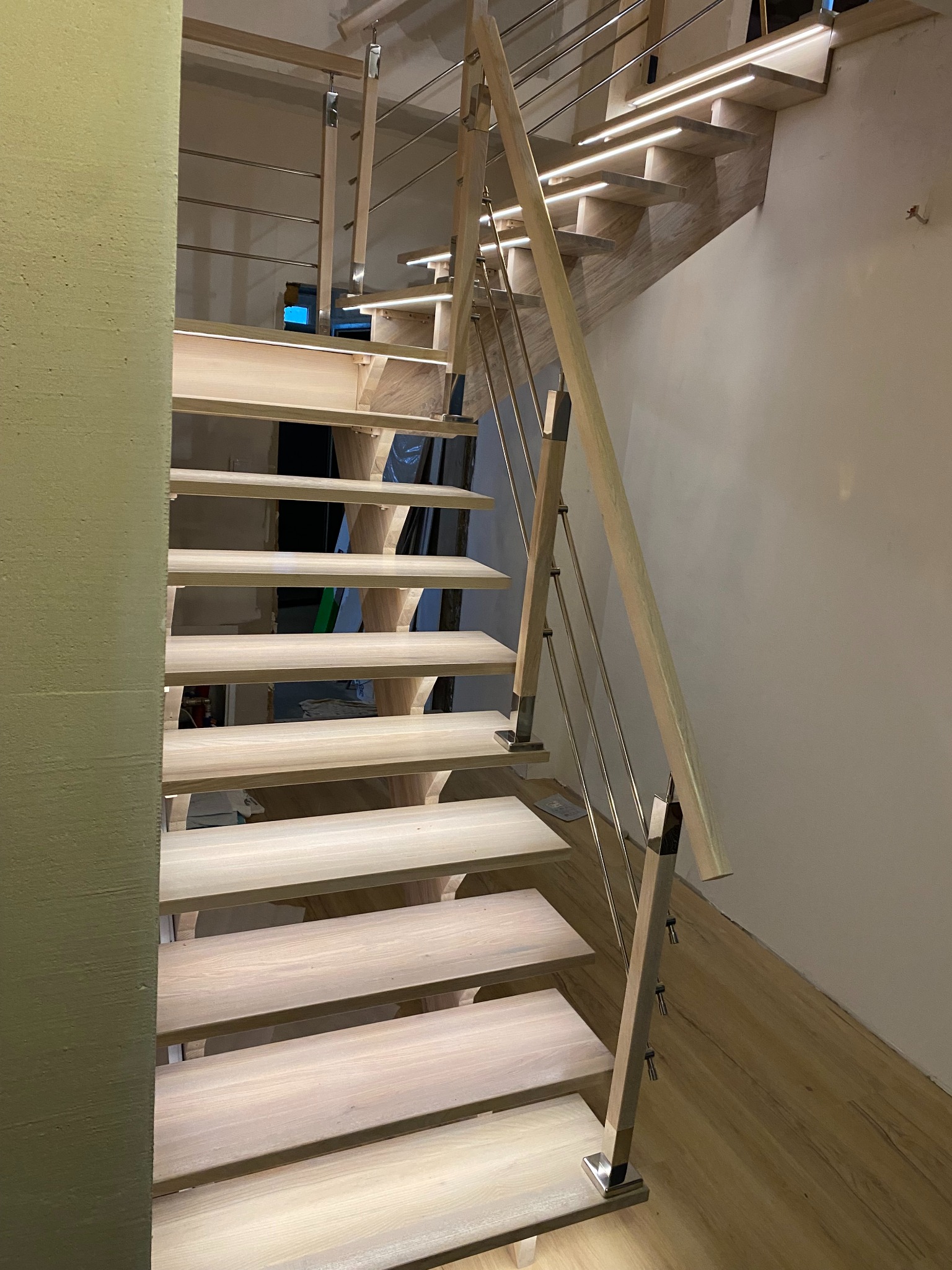 Nowoczesne schody z jasnego drewna z metalowymi elementami i subtelnym oświetleniem LED pod stopniami, widok z dołu, ukazujący detale konstrukcji i wykończenia.