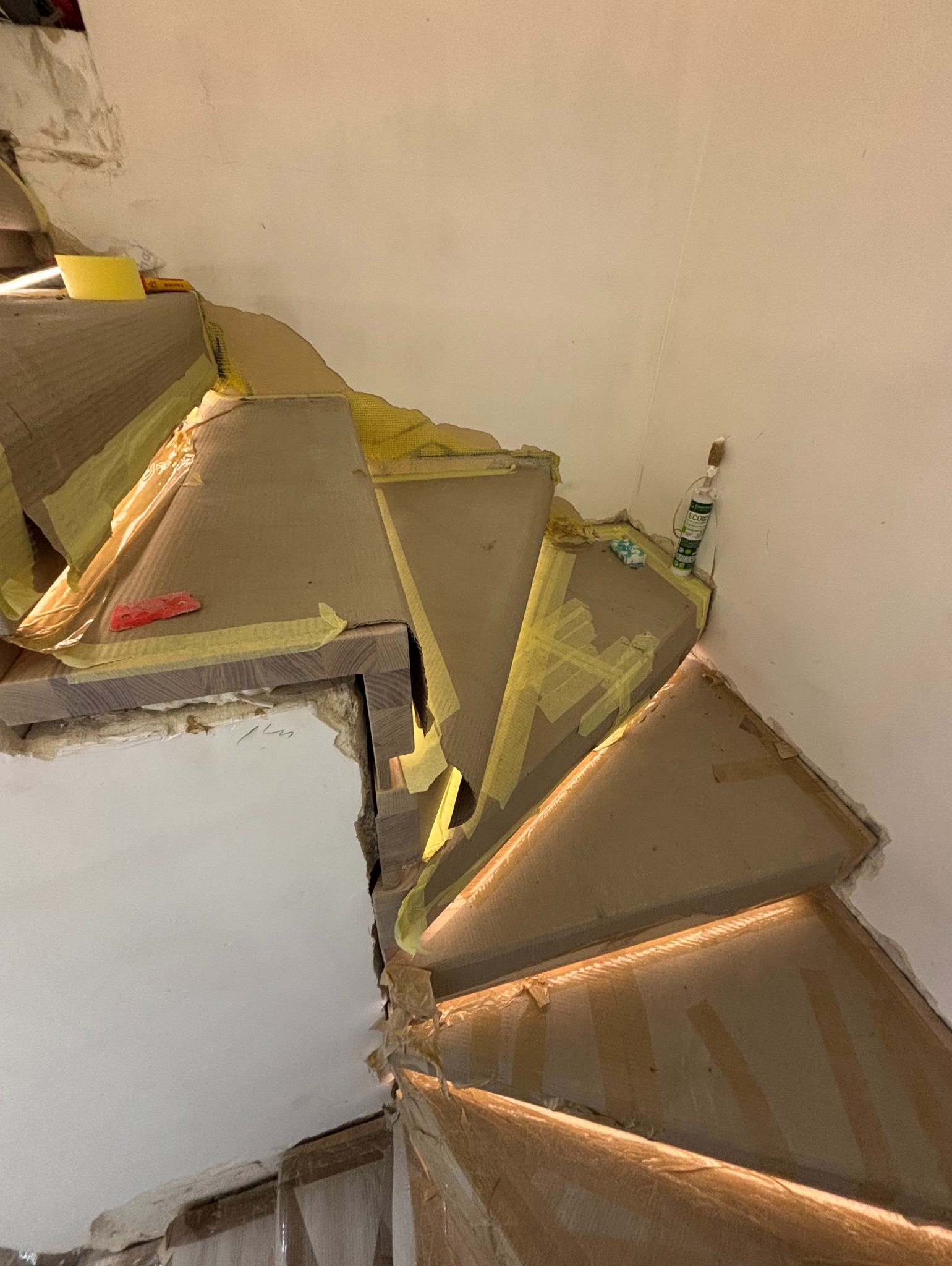 Montaż drewnianych schodów z podświetleniem LED, zabezpieczonych kartonem i taśmą malarską. Widoczne narzędzia i materiały montażowe. Ujęcie z góry, w trakcie prac wykończeniowych.