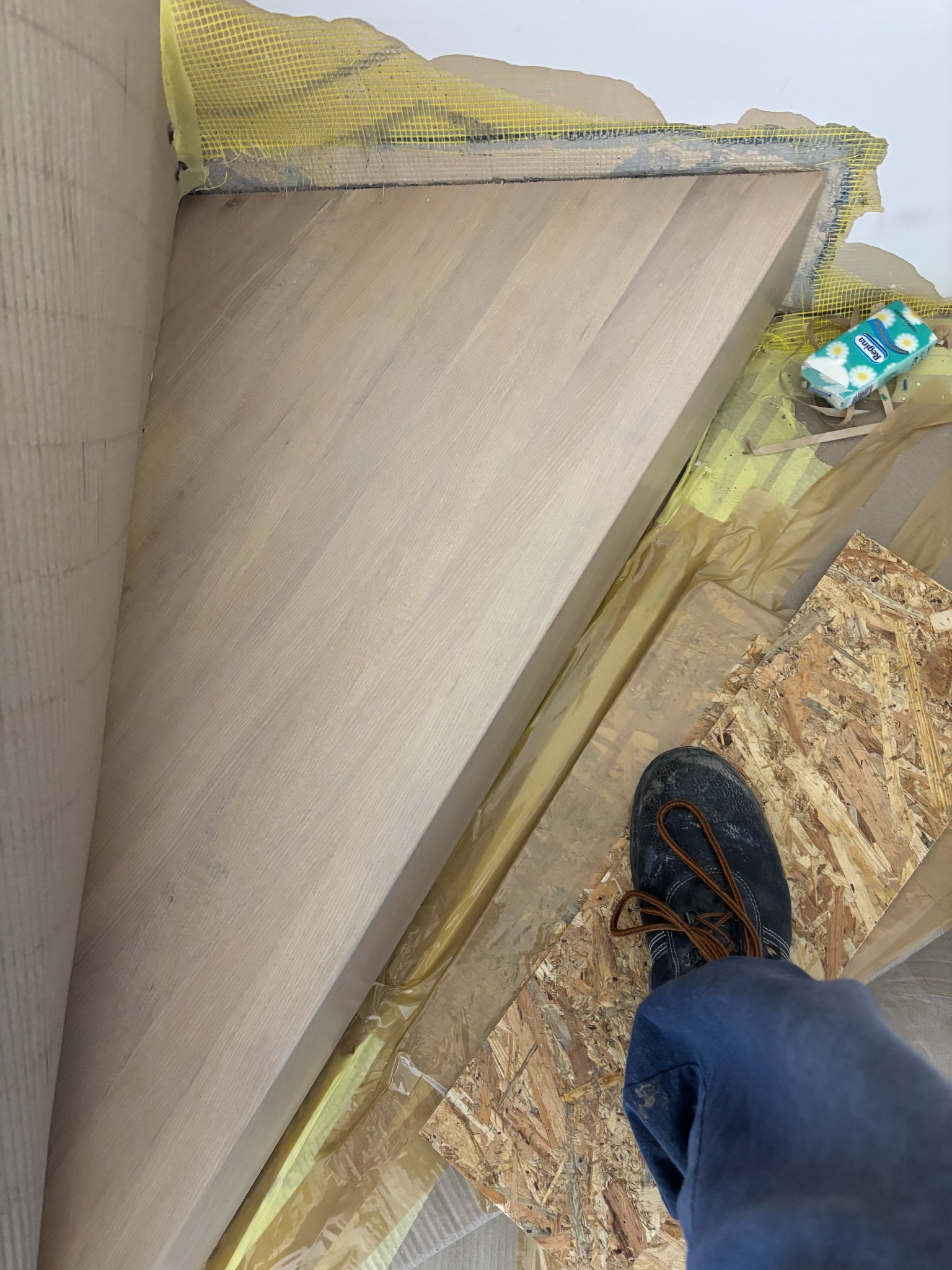 Drewniane schody w trakcie montażu, widok z góry na stopień z fragmentem buta pracownika. Żółta siatka zabezpieczająca na ścianie i folia ochronna na podłodze.