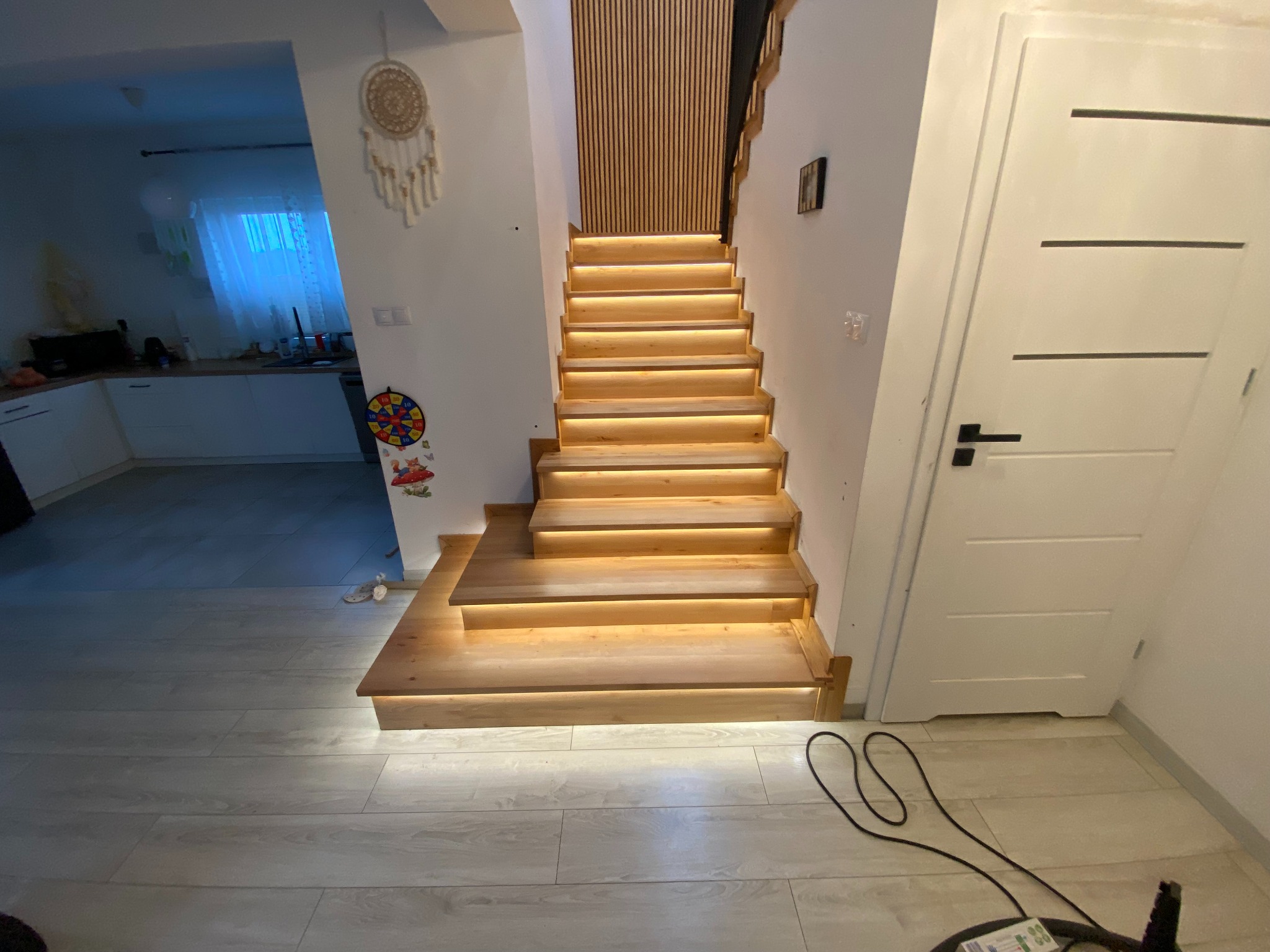 Podświetlane, jasne schody drewniane w nowoczesnym wnętrzu. Widoczne listwy dekoracyjne na ścianie i fragment kuchni w tle. Ujęcie z dołu, podkreślające detale wykończenia.