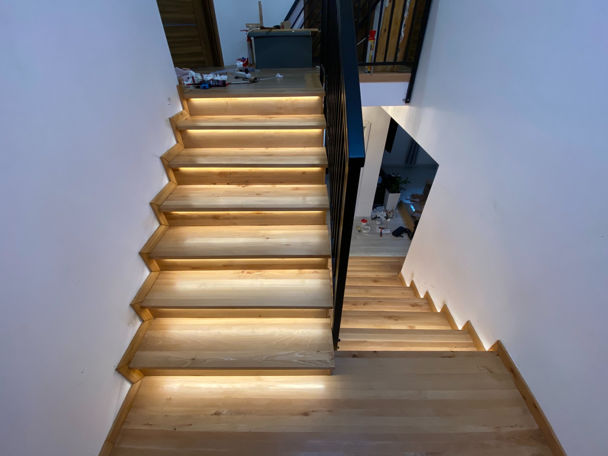 Nowoczesne schody z jasnego drewna z podświetleniem LED, widok z góry. Minimalistyczna balustrada z czarnych prętów. Białe ściany. Widoczne elementy wykończenia wnętrza.