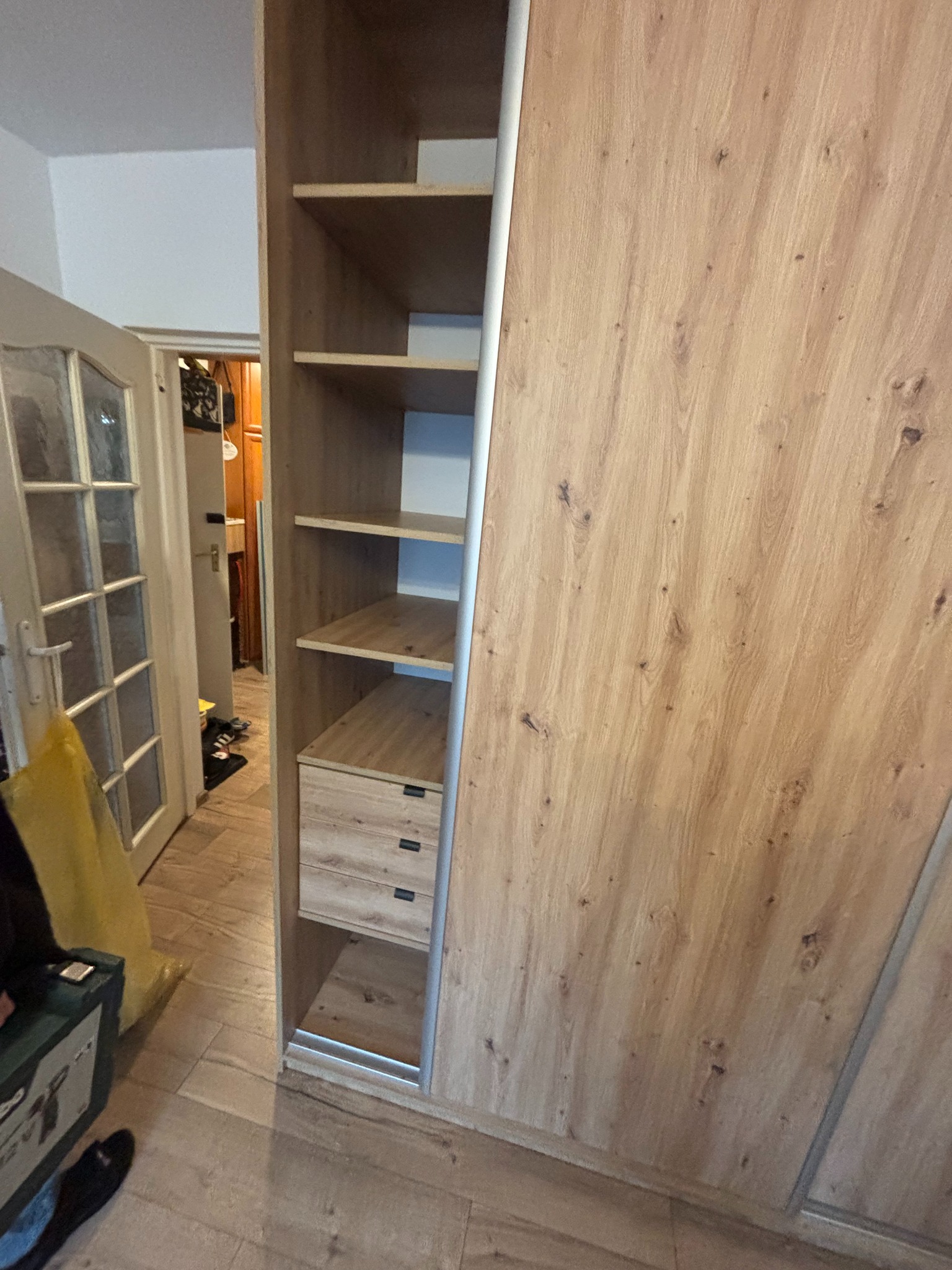 Jasna, drewnopodobna szafa wnękowa z przesuwnymi drzwiami i otwartymi półkami, widoczna z boku, z perspektywy podłogi. W tle drzwi do innego pomieszczenia.